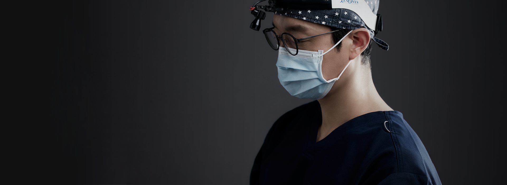 수술 모자·헤드라이트·확대경 안경·마스크를 쓴 집도의 옆모습(어두운 바탕)